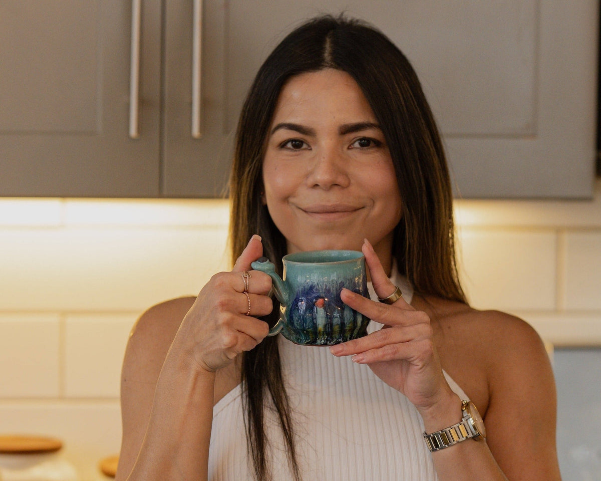 A woman holding a blue and green textured ceramic mug in a kitchen setting with beige cabinets and tiled backsplash.