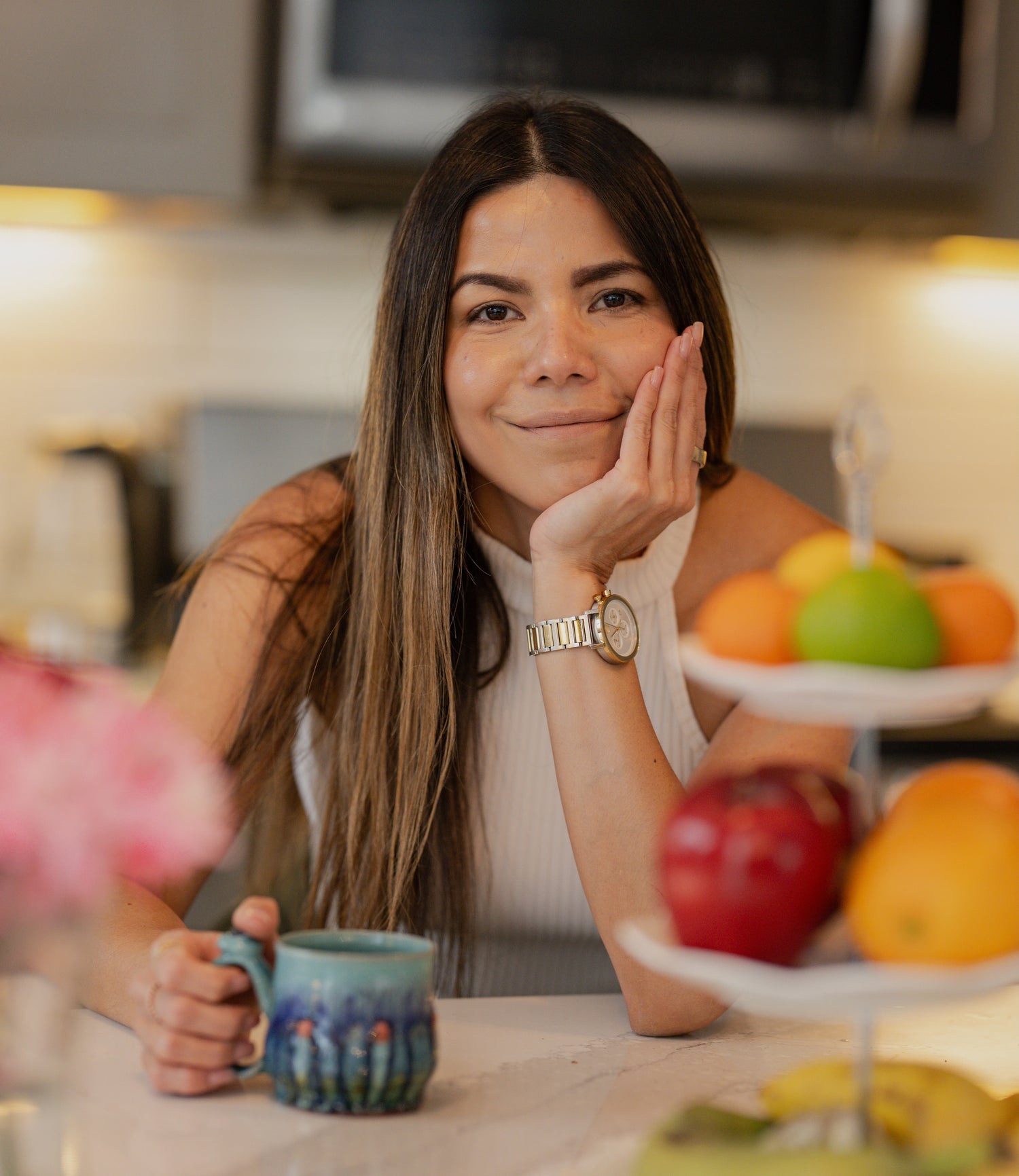 Woman sitting at a kitchen table with a mug, surrounded by fruits and a cake stand.